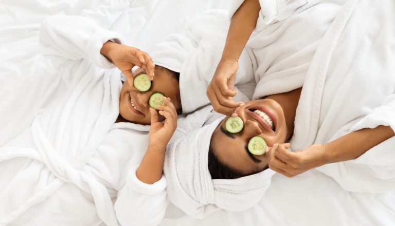 A mother and daughter, both wearing white robes and head towels, relaxing together during an at-home spa day with cucumber slices over their eyes.