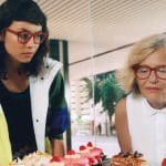 A young woman and an older woman, each wearing a pair of glasses with large red frames. They are standing over some pastries.