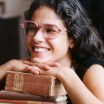 A smiling woman wearing a pair of glasses with large pink frames, resting her chin on her folded hands, on top of a stack of books.