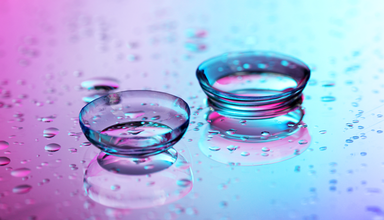 A close up picture of two contact lenses on a purple and blue background, surrounded by drops of water.