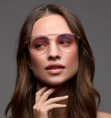 Model wearing new arrival Shamir PhotoGlamour™ coloured Transitions® Light Intelligent Lenses™ in pink, posing with a hand on their neck and a curious, confident expression.