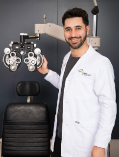 Dr. Jordan Kassouf stands in front of eye exam equipment, wearing a white lab coat over a grey professional shirt. He has black hair and neatly groomed facial hair.