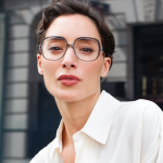 A woman wearing Lafont wide-style frames and a white blouse poses outside against a grey brick building.