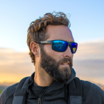 A man in tactical gear wearing Wiley X eyewear stands outdoors at sunset, looking serious.